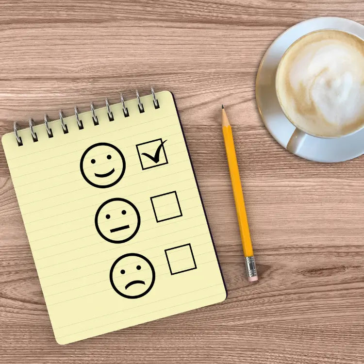 Carnet à spirales posé sur une table en bois, montrant trois visages dessinés : un visage souriant avec une case cochée, un visage neutre et un visage triste avec leurs cases vides. Un crayon jaune est posé à côté et, en haut à droite, se trouve une tasse de café avec mousse.