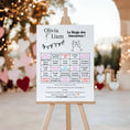 Charger l'image dans la visionneuse de la galerie, Affiche grand format du jeu “Bingo des Amoureux” personnalisée aux prénoms, présentée sur un chevalet lors d’un mariage en extérieur. Le décor romantique comporte des guirlandes lumineuses, des cœurs et un fond flou festif.