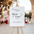 Panneau de bienvenue mariage personnalisé avec prénoms et date. Exemple Olivia et Liam, affiche élégante sur chevalet décoré de cœurs et guirlandes lumineuses.