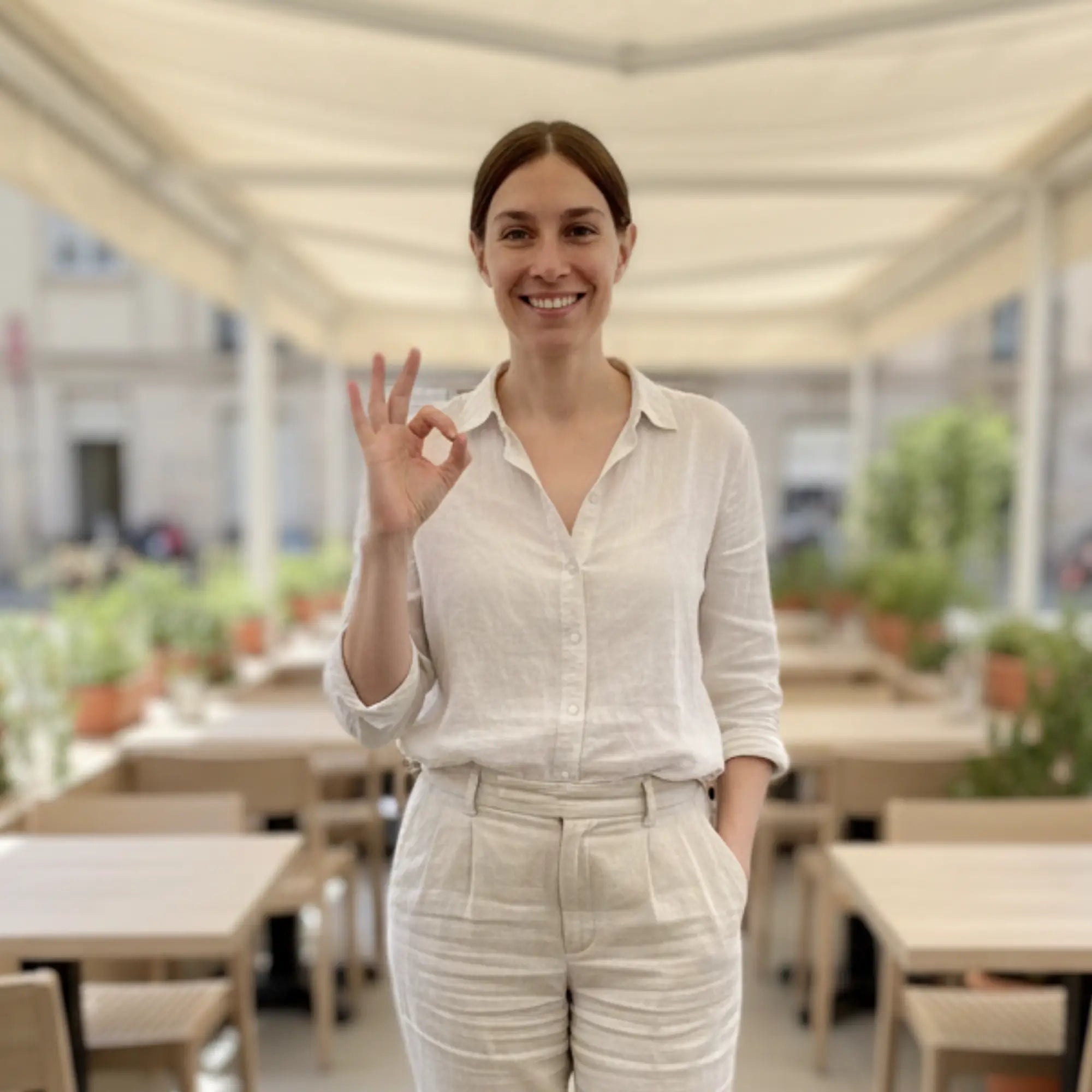 Femme souriante dans un café en plein air, faisant un signe OK, illustrant la présentation de Template Boutique Cie et de ses modèles Canva personnalisables pour mariage, baptême, naissance et événements.