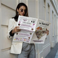 Charger l'image dans la visionneuse de la galerie, Une femme en tailleur blanc lit la Gazette EVJF Love. La page gauche présente la playlist spéciale, le jeu '2 vérités, 1 mensonge' et les 3 cocktails EVJF, tandis que la couverture à droite affiche un grand cœur composé de photos souvenirs et la phrase 'Un week-end, des copines, des fous rires… et des secrets à garder à vie'
