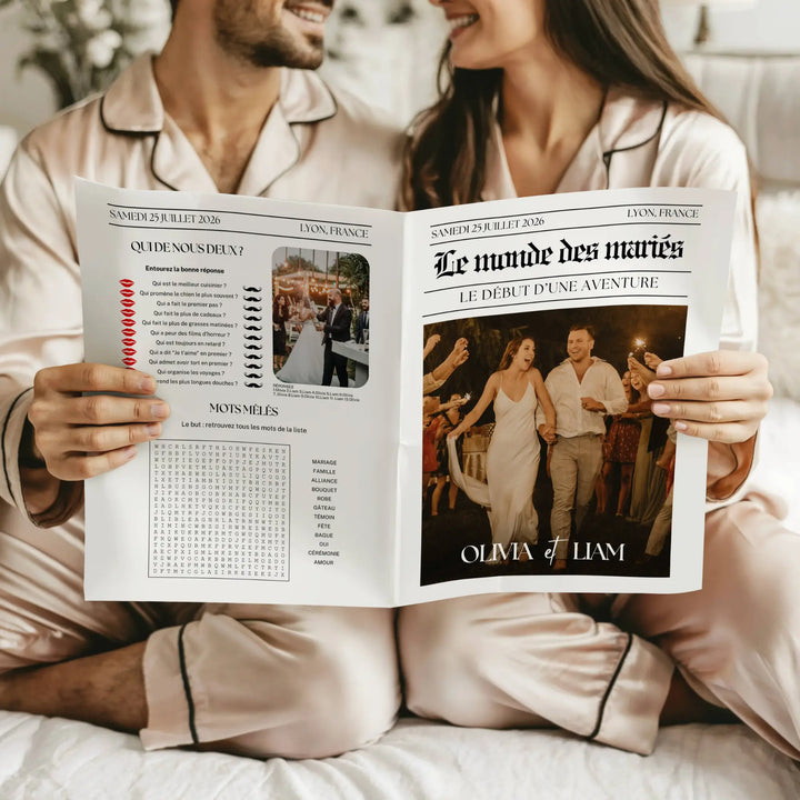 Couple en pyjama satiné beige assis côte à côte, tenant ensemble une gazette de mariage bohème, présentant en couverture les mariés marchant main dans la main entourés d’invités, et à gauche un quiz humoristique avec une grille de mots mêlés sur le thème du mariage.