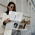 Charger l'image dans la visionneuse de la galerie, Femme élégante portant un blazer blanc et des lunettes de soleil rondes, lisant une gazette de mariage originale. La scène se déroule dans une rue lumineuse au style urbain chic.
