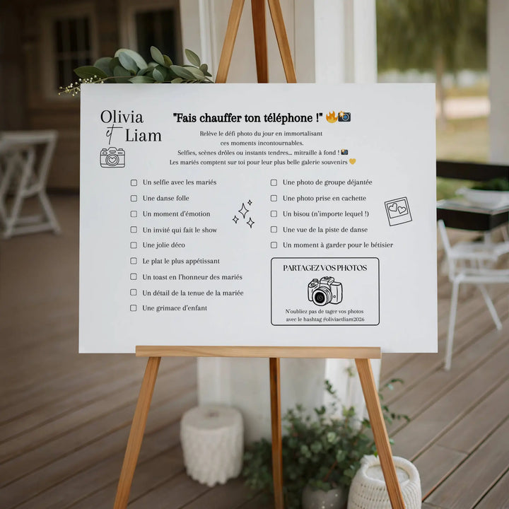 Affiche de jeu mariage « Fais chauffer ton téléphone ! » sur chevalet en bois, invitant les invités à relever des défis photos (selfie avec les mariés, danse folle, décor, photo de groupe…) avec cadre élégant et zone “Partagez vos photos” avec hashtag personnalisé.