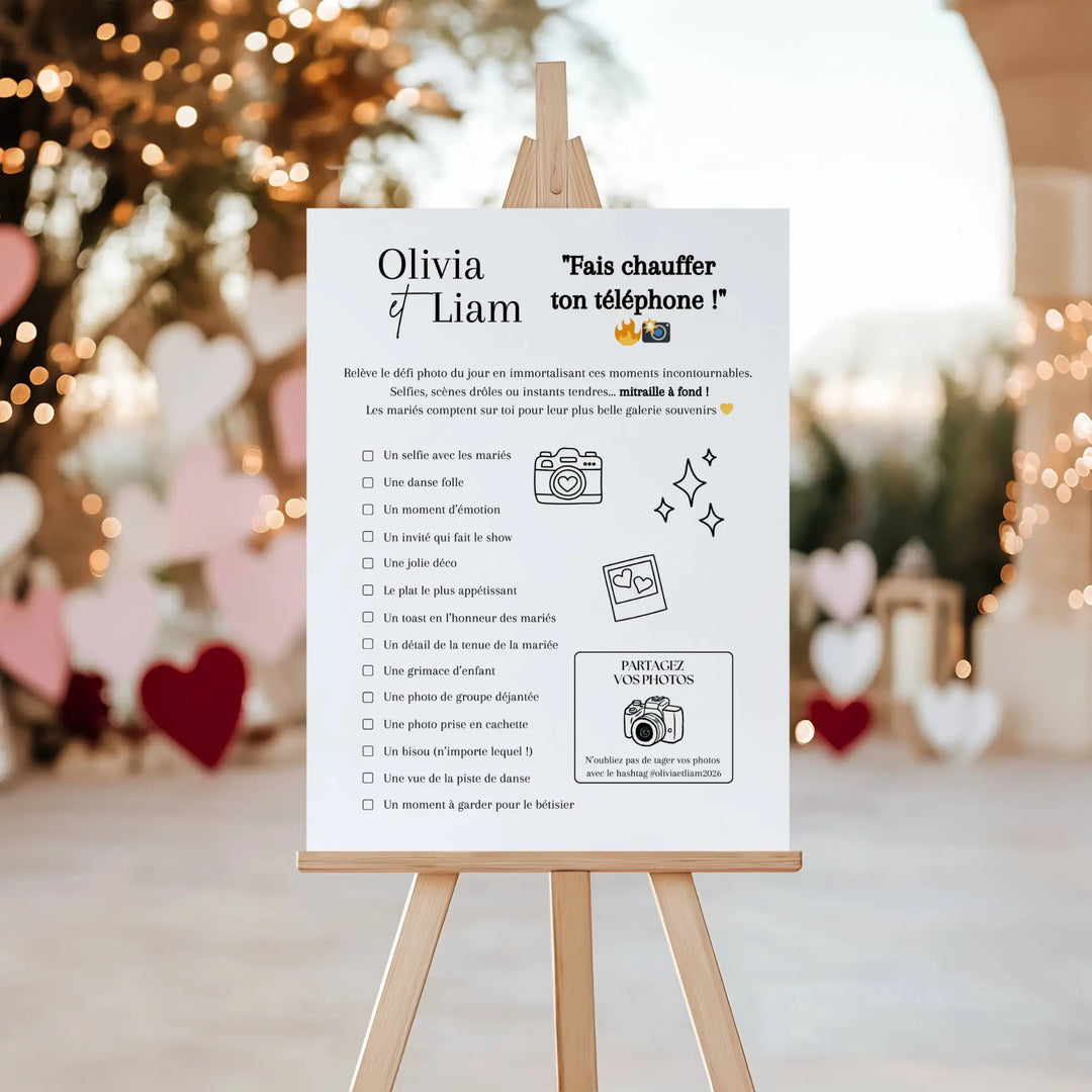 Affiche de jeu de mariage « Fais chauffer ton téléphone ! » posée sur un chevalet en bois, devant une décoration romantique avec guirlandes lumineuses et cœurs, invitant les invités à relever des défis photos et à partager leurs clichés des mariés.