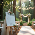Charger l'image dans la visionneuse de la galerie, Affiche de jeu labyrinthe de mariage personnalisée sur chevalet en bois, décorée de fleurs, installée dans une allée de cérémonie en forêt avec arche fleurie en arrière-plan et guirlande lumineuse suspendue.
