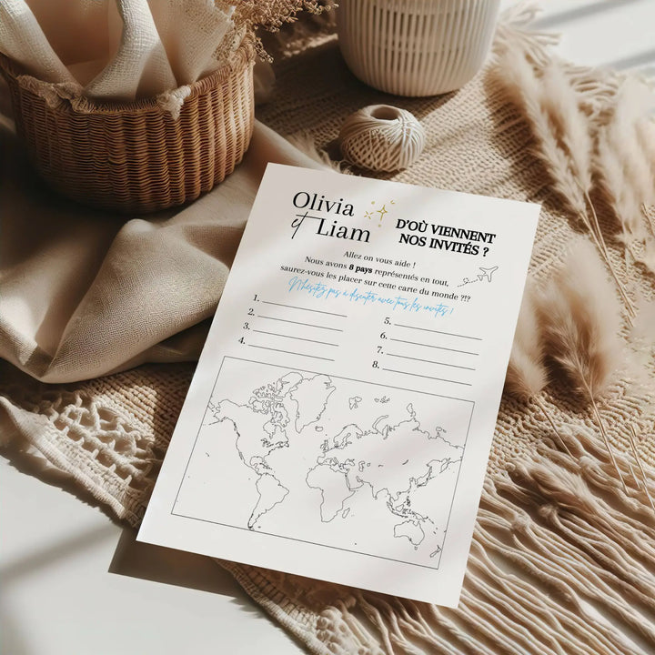 Fiche de jeu de mariage “D’où viennent nos invités ?” posée sur un plaid beige, entourée d’éléments bohèmes (panier en osier, pampas), avec carte du monde à remplir.