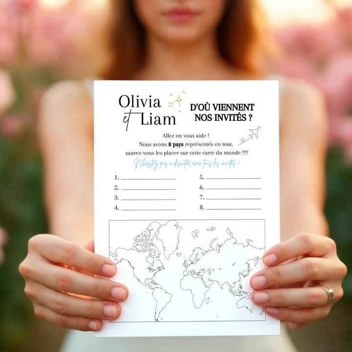 Jeu de mariage “D’où viennent nos invités ?” tenu à deux mains devant un décor fleuri, avec carte du monde et emplacements à remplir pour deviner les pays des invités.