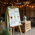 Charger l'image dans la visionneuse de la galerie, Panneau de menu bar de mariage sur chevalet en bois, décoré d’un tissu vert. Affiche personnalisée avec cocktails signatures (Americano, Mojito, Mocktail), champagnes, vins, bières et boissons sans alcool. Décoration élégante pour bar de mariage.
