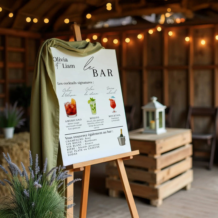 Panneau de menu bar de mariage sur chevalet en bois, décoré d’un tissu vert. Affiche personnalisée avec cocktails signatures (Americano, Mojito, Mocktail), champagnes, vins, bières et boissons sans alcool. Décoration élégante pour bar de mariage.