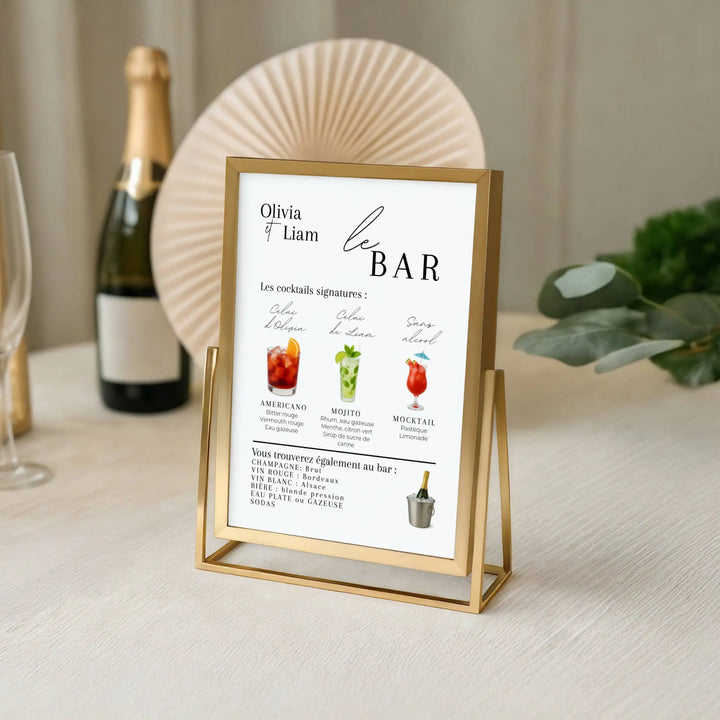 Menu bar de mariage encadré en doré posé sur une table élégante. Affiche personnalisée avec cocktails signatures (Americano, Mojito, Mocktail), champagnes, vins, bières et boissons sans alcool. Décoration chic et moderne pour table de mariage.