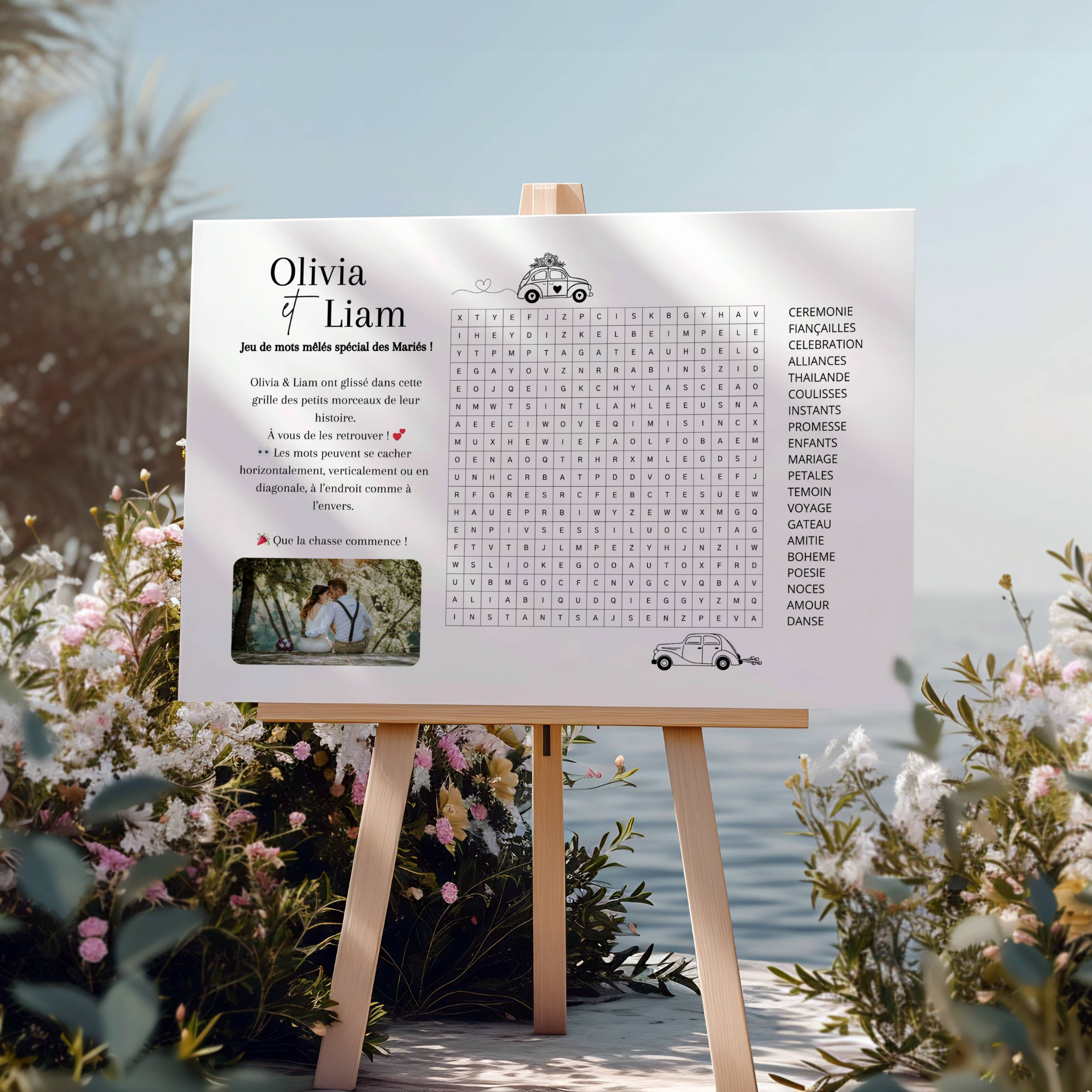 Affiche de jeu de mots mêlés personnalisée au nom des mariés, présentée sur un chevalet en bois dans un décor fleuri au bord de l’eau. Au centre se trouve une grande grille de mots mêlés, avec à droite la liste des mots à retrouver (comme “cérémonie”, “amour”, “voyage”, “témoin”…). À gauche, un petit texte explicatif invite les invités à jouer, accompagné d’illustrations de voiture de mariage. En bas à gauche se trouve une photo du couple. L’ambiance est romantique et champêtre.
