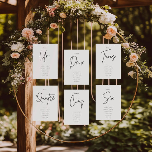 Plan de table mariage avec numéros élégants – cartes minimalistes suspendues dans une arche fleurie.