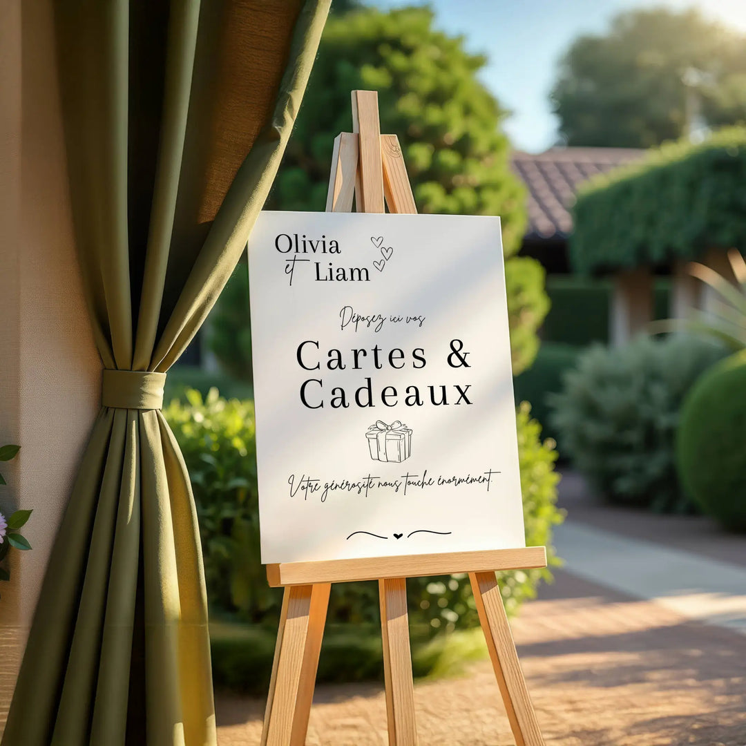 Panneau de mariage “Cartes & Cadeaux” personnalisé avec les prénoms Olivia & Liam, installé sur un chevalet en bois à l’extérieur. Texte élégant invitant les invités à déposer leurs cartes et cadeaux, décor raffiné avec rideau vert et jardin en arrière-plan.