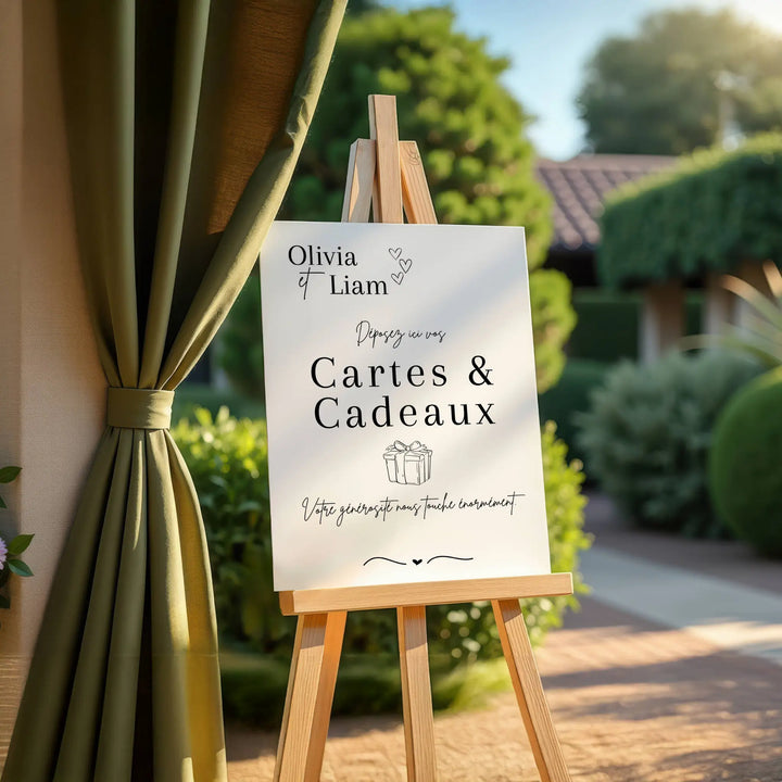 Panneau de mariage “Cartes & Cadeaux” personnalisé avec les prénoms Olivia & Liam, installé sur un chevalet en bois à l’extérieur. Texte élégant invitant les invités à déposer leurs cartes et cadeaux, décor raffiné avec rideau vert et jardin en arrière-plan.
