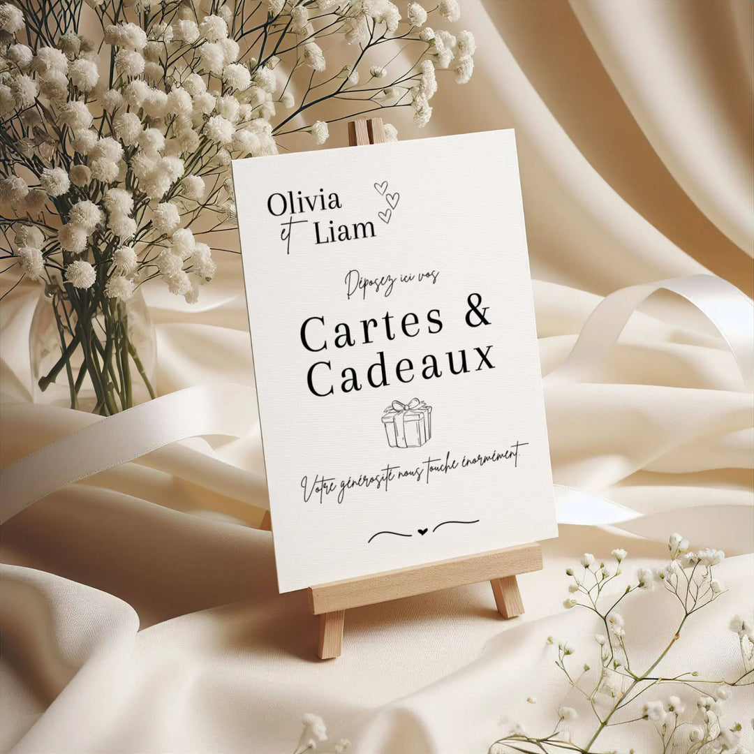 Panneau de mariage personnalisé “Cartes & Cadeaux” au nom d’Olivia & Liam, imprimé sur papier texturé et présenté sur un petit chevalet en bois, entouré de fleurs de gypsophile et de tissu ivoire élégant.