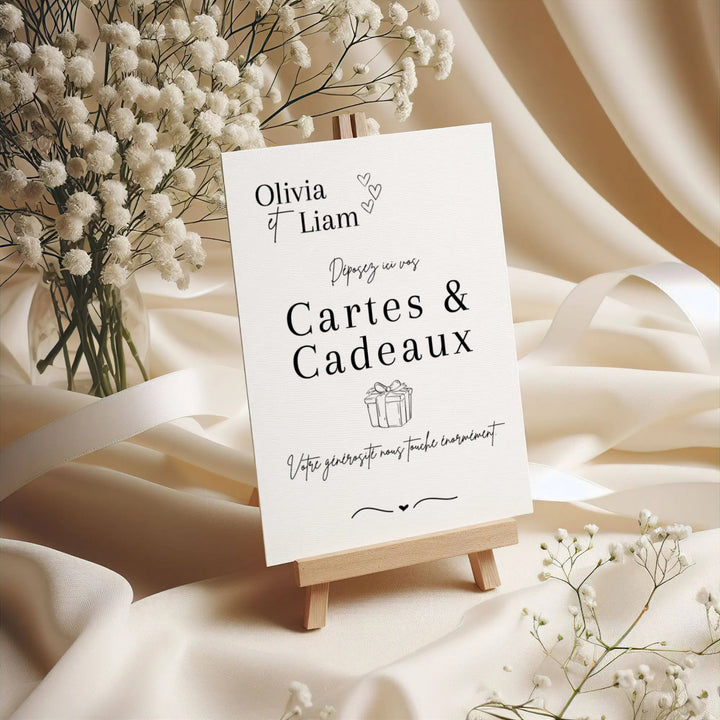 Panneau de mariage personnalisé “Cartes & Cadeaux” au nom d’Olivia & Liam, imprimé sur papier texturé et présenté sur un petit chevalet en bois, entouré de fleurs de gypsophile et de tissu ivoire élégant.