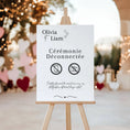 Charger l'image dans la visionneuse de la galerie, Panneau de mariage personnalisé “Cérémonie Déconnectée” avec prénoms Olivia et Liam, design élégant en noir et blanc avec icônes téléphone et appareil photo barrés. Le panneau est présenté sur un chevalet en bois, décor romantique avec lumières et cœurs en arrière-plan. Message : profitez pleinement du moment, nos photographes capturent chaque instant.
