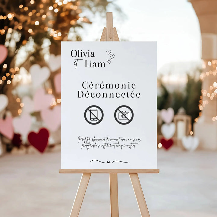 Panneau de mariage personnalisé “Cérémonie Déconnectée” avec prénoms Olivia et Liam, design élégant en noir et blanc avec icônes téléphone et appareil photo barrés. Le panneau est présenté sur un chevalet en bois, décor romantique avec lumières et cœurs en arrière-plan. Message : profitez pleinement du moment, nos photographes capturent chaque instant.