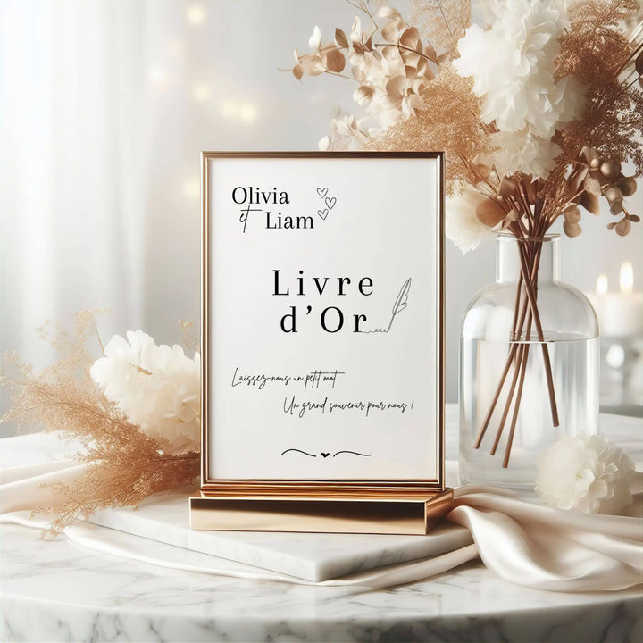 Panneau de mariage “Livre d’or” personnalisé au nom d’Olivia & Liam, présenté dans un cadre doré élégant sur une table décorée de fleurs blanches et dorées. Texte invitant les invités à laisser un petit mot souvenir.