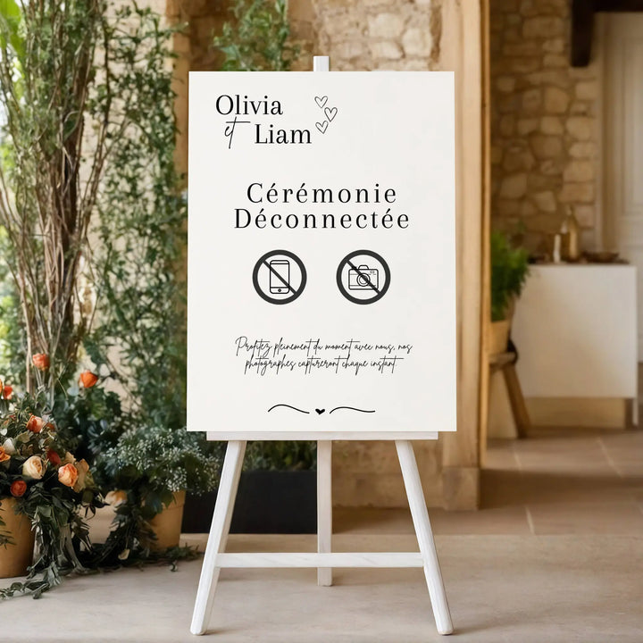 Panneau de mariage “Cérémonie Déconnectée” personnalisé avec les prénoms Olivia & Liam, installé sur un chevalet blanc dans une salle en pierre décorée de fleurs. Icônes téléphone et appareil photo barrés invitant les invités à profiter du moment sans écrans.